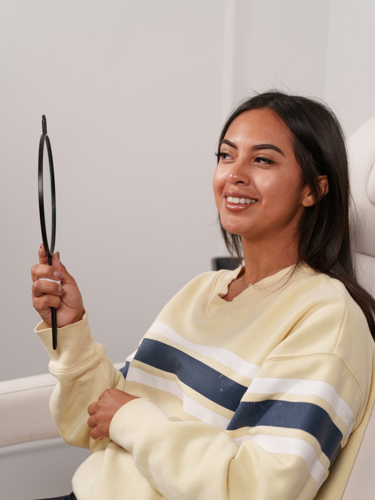 A smiling woman looking at her skin in a hand mirror in a treatment room